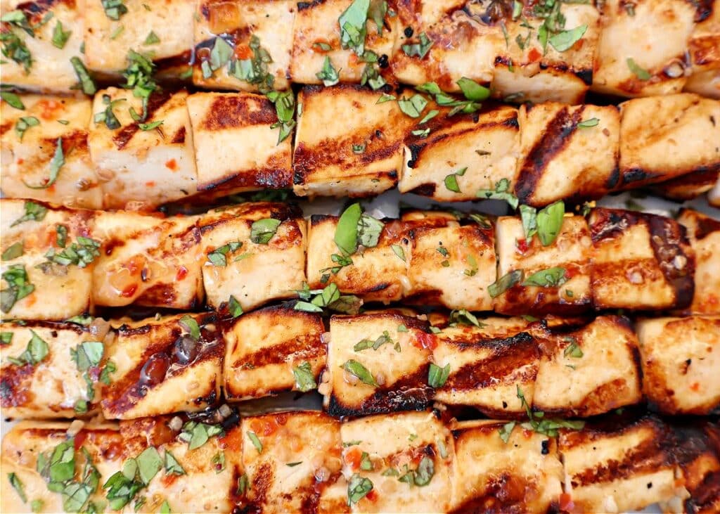 Grilled tofu skewers garnished with chopped fresh herbs, arranged closely together. The paneer cubes have charred grill marks and a coating of spices and herbs.