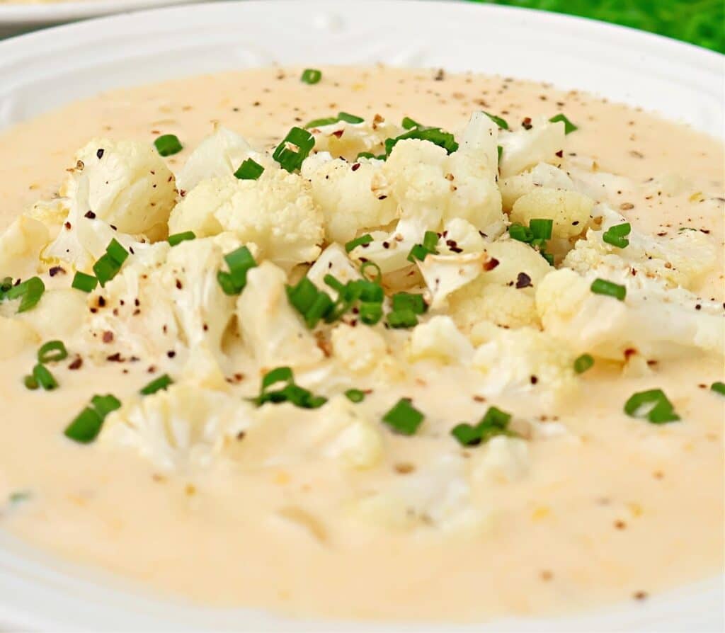 A bowl of creamy cauliflower soup topped with chopped chives and freshly ground black pepper, served on a white plate.