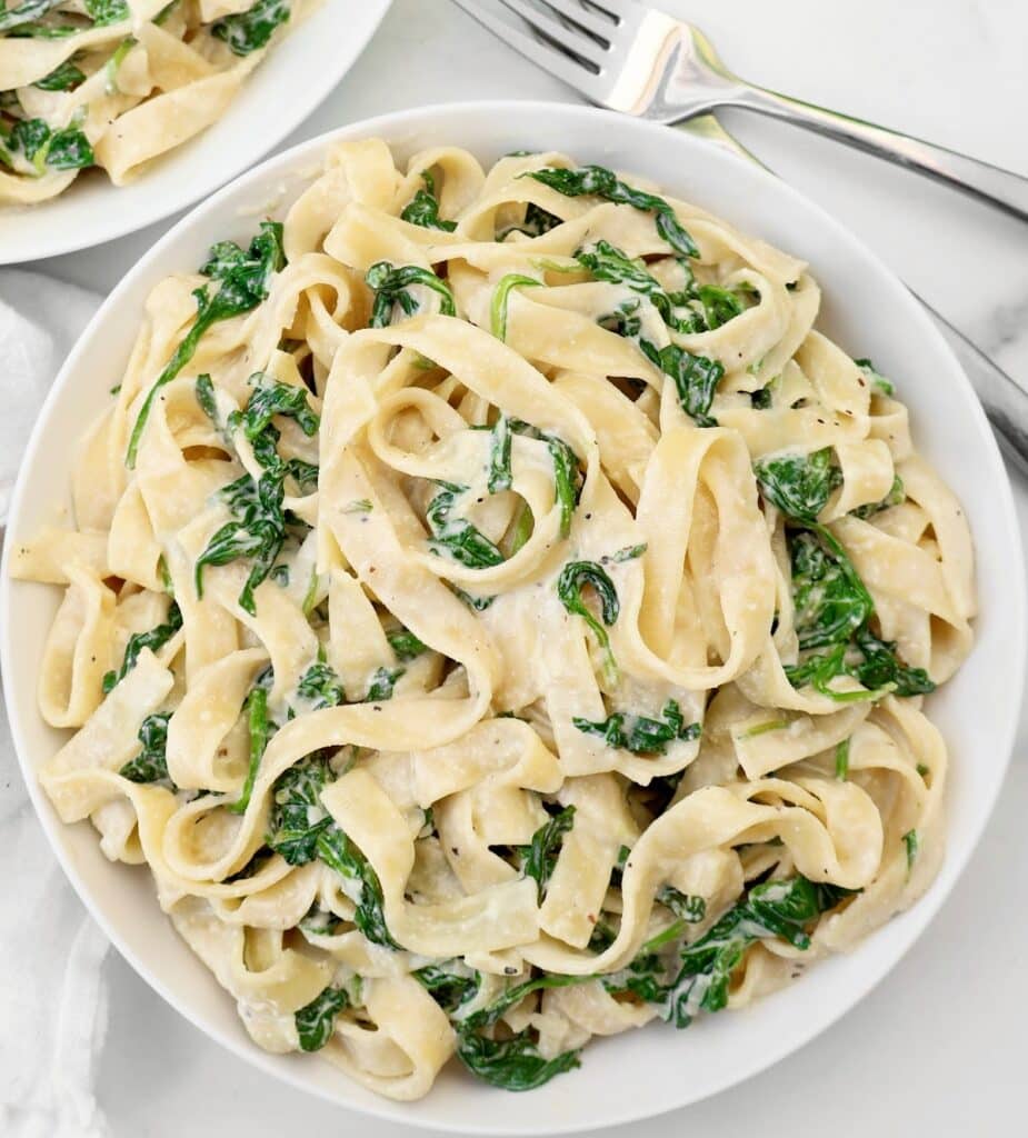 A bowl of creamy fettuccine pasta mixed with sautéed spinach, served on a white plate with a fork nearby on a white marble surface.