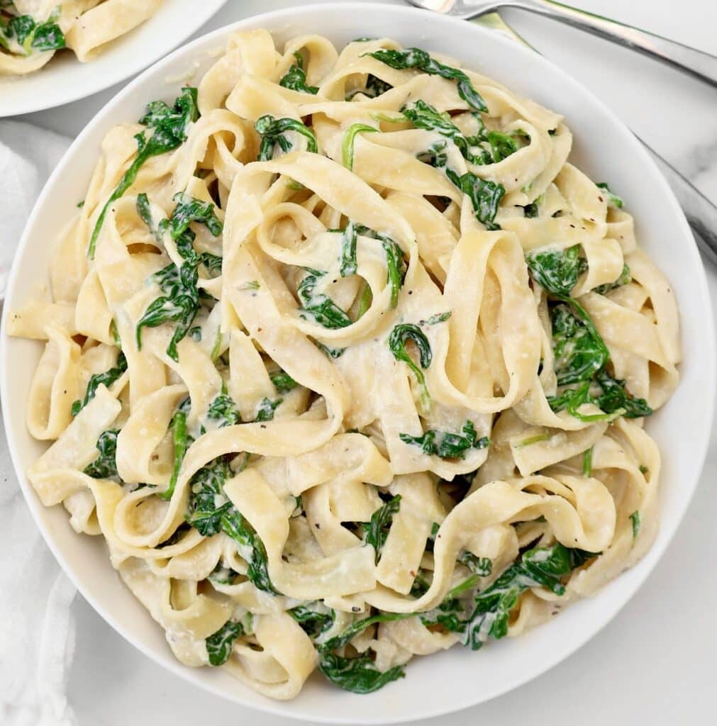 A white bowl filled with creamy fettuccine pasta mixed with wilted spinach, placed on a white marble surface. Fork and knife are partially visible at the top of the image.