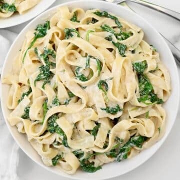 A white bowl filled with creamy fettuccine pasta mixed with wilted spinach, placed on a white marble surface. Fork and knife are partially visible at the top of the image.