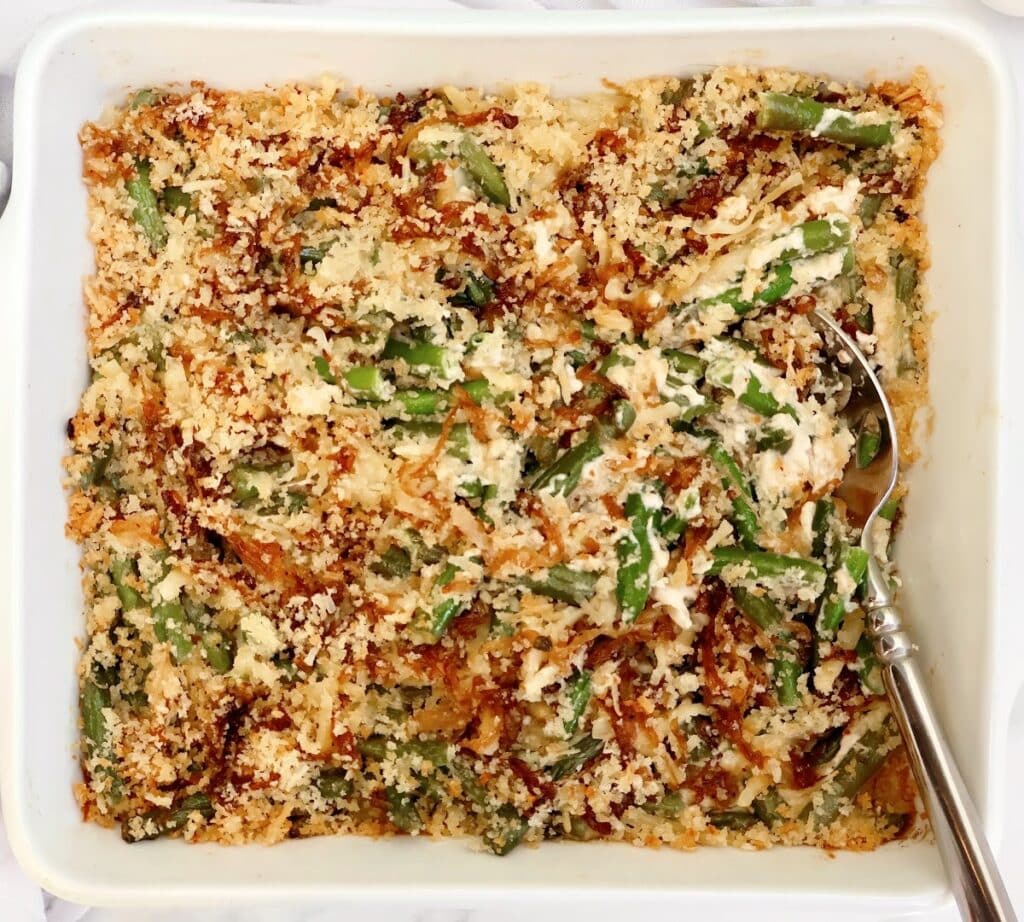 A baked green bean casserole topped with crispy, golden breadcrumbs, served in a white dish with a silver serving spoon on the right side.