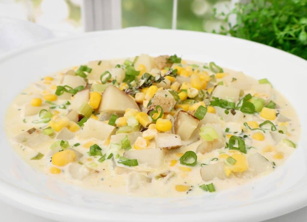 A bowl of creamy corn chowder topped with diced potatoes, corn kernels, and chopped green onions, garnished with fresh herbs, served on a white plate with greenery in the background.