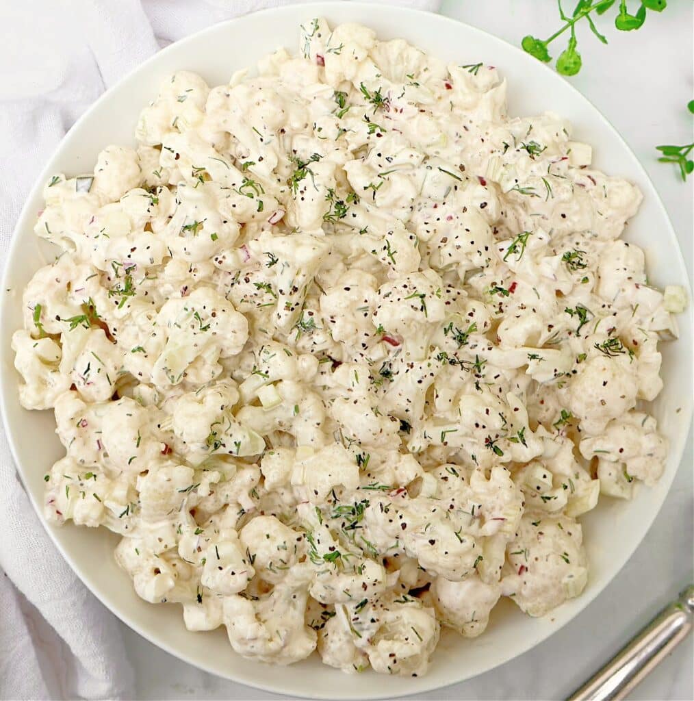 A white bowl filled with creamy cauliflower salad, garnished with chopped herbs and cracked black pepper, on a white surface with a napkin and silver utensil beside it.