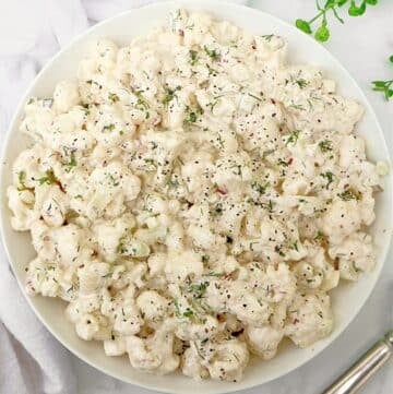 A white bowl filled with creamy cauliflower salad, garnished with chopped herbs and cracked black pepper, on a white surface with a napkin and silver utensil beside it.