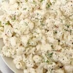 A close-up of a creamy cauliflower salad with chopped red onions and herbs, all coated in a white dressing and garnished with fresh dill on a white plate.