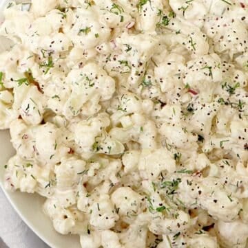 A close-up of a creamy cauliflower salad with chopped red onions and herbs, all coated in a white dressing and garnished with fresh dill on a white plate.