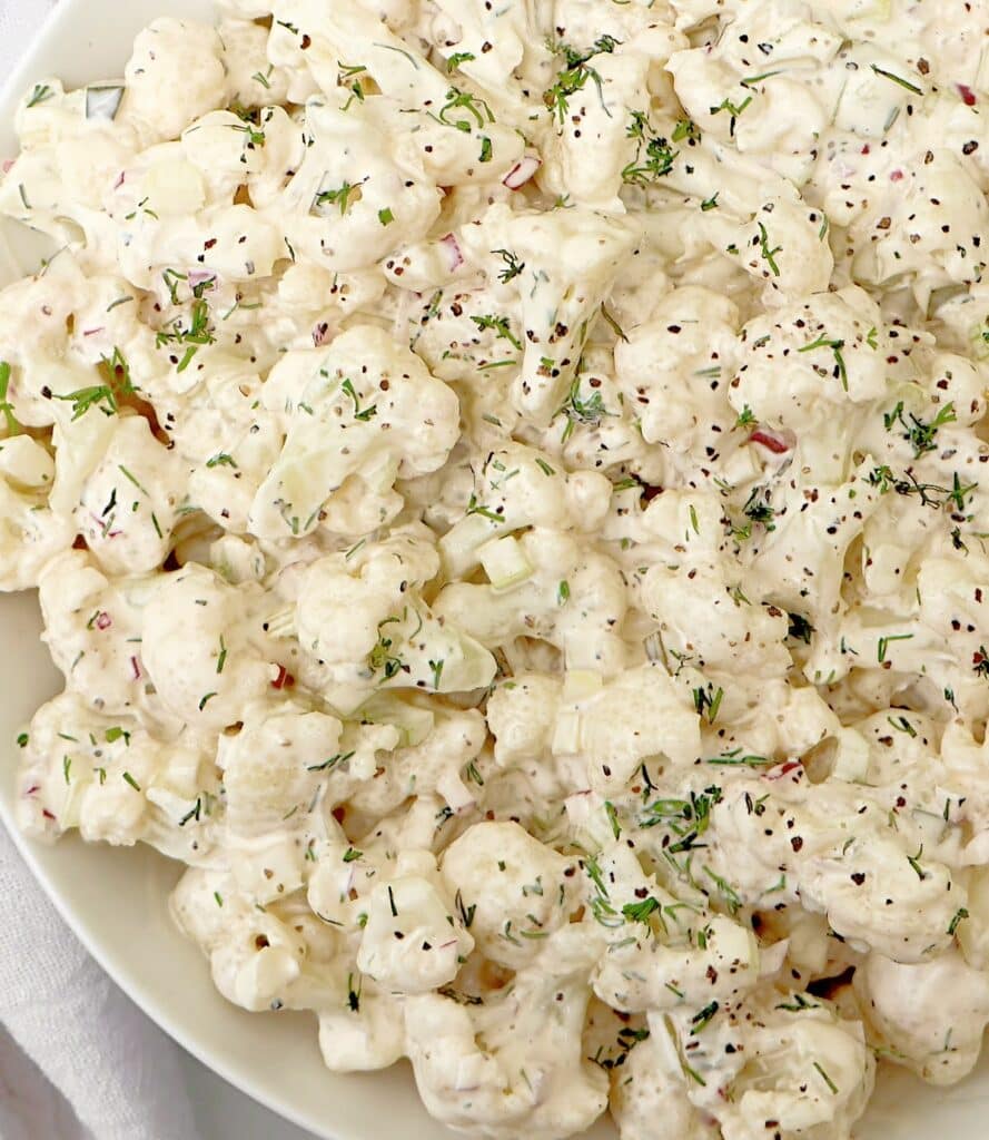 A close-up of a creamy cauliflower salad with chopped red onions and herbs, all coated in a white dressing and garnished with fresh dill on a white plate.