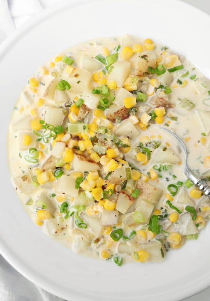 A creamy soup with diced potatoes, corn kernels, chopped green onions, and herbs, served in a white bowl with a spoon.