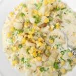 A close-up of a creamy corn chowder soup with diced potatoes, corn kernels, chopped green onions, and celery, served in a white bowl with a fork.
