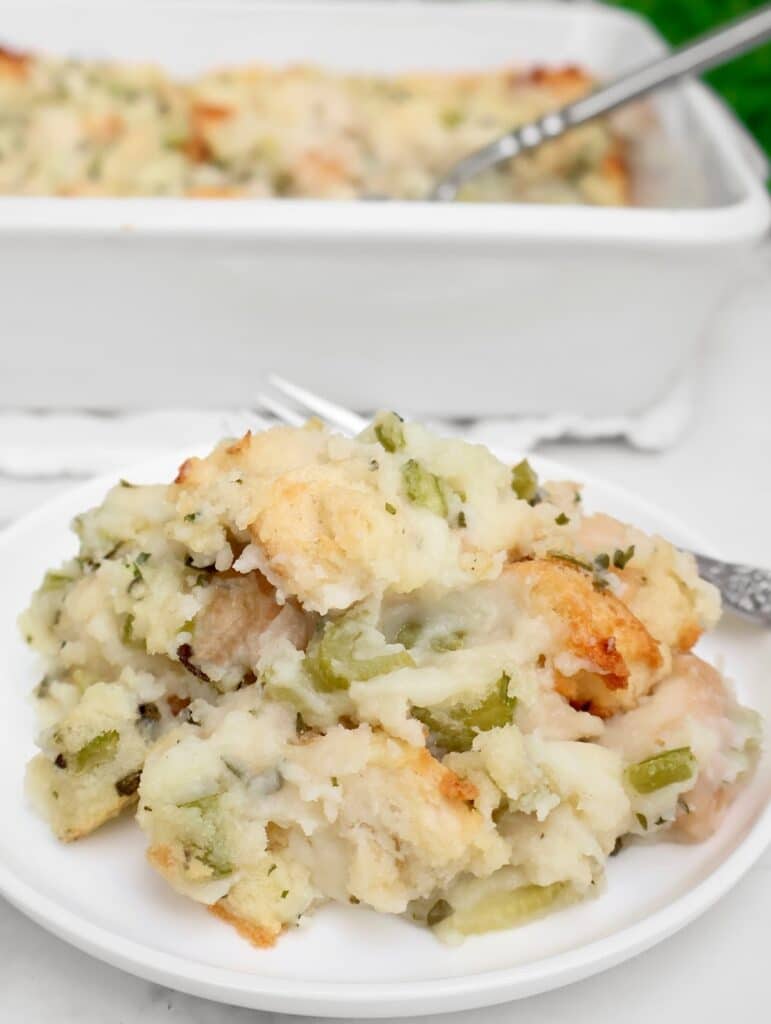 A serving of baked bread stuffing with celery and herbs is on a white plate, with a fork beside it. In the background, a white baking dish filled with more stuffing is visible.