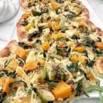 A flatbread topped with roasted vegetables including butternut squash, Brussels sprouts, leafy greens, and shredded cheese, displayed on a white surface with green herbs in the background.