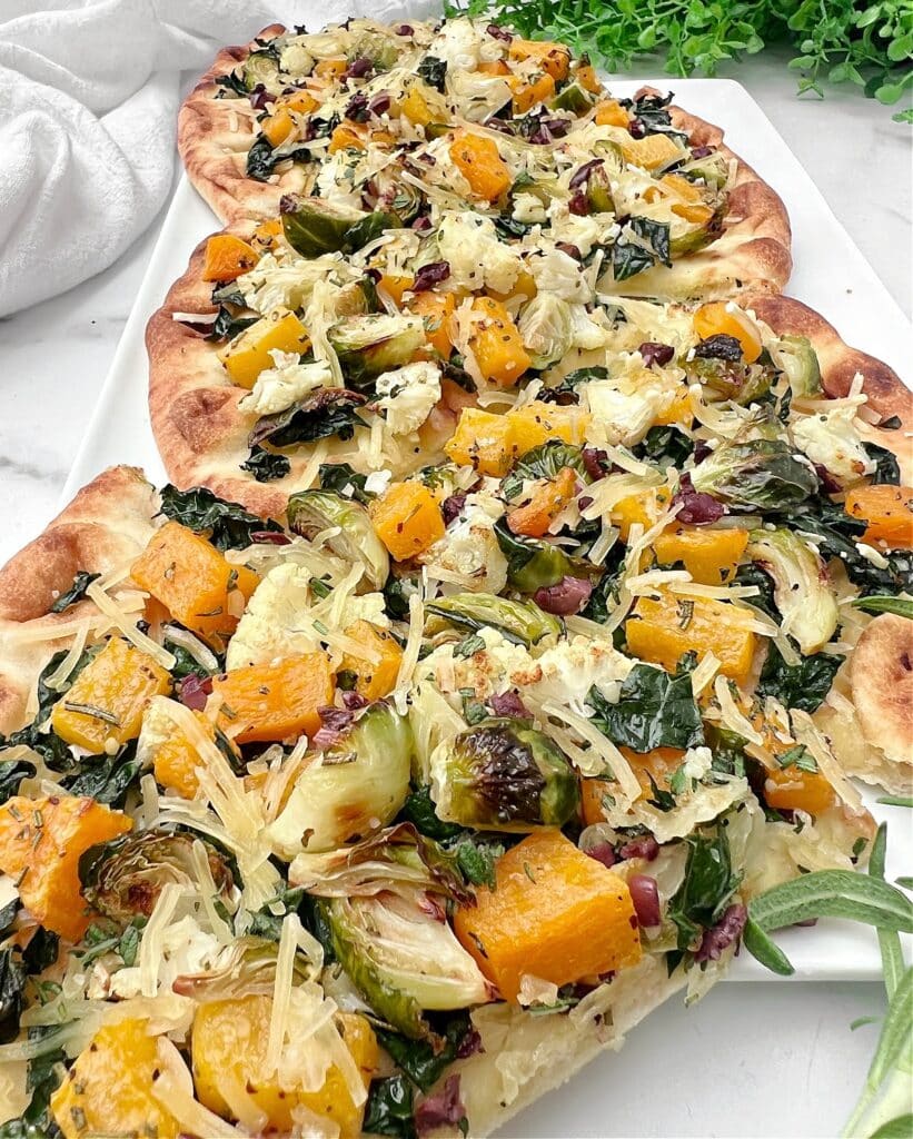 A flatbread topped with roasted vegetables including butternut squash, Brussels sprouts, leafy greens, and shredded cheese, displayed on a white surface with green herbs in the background.