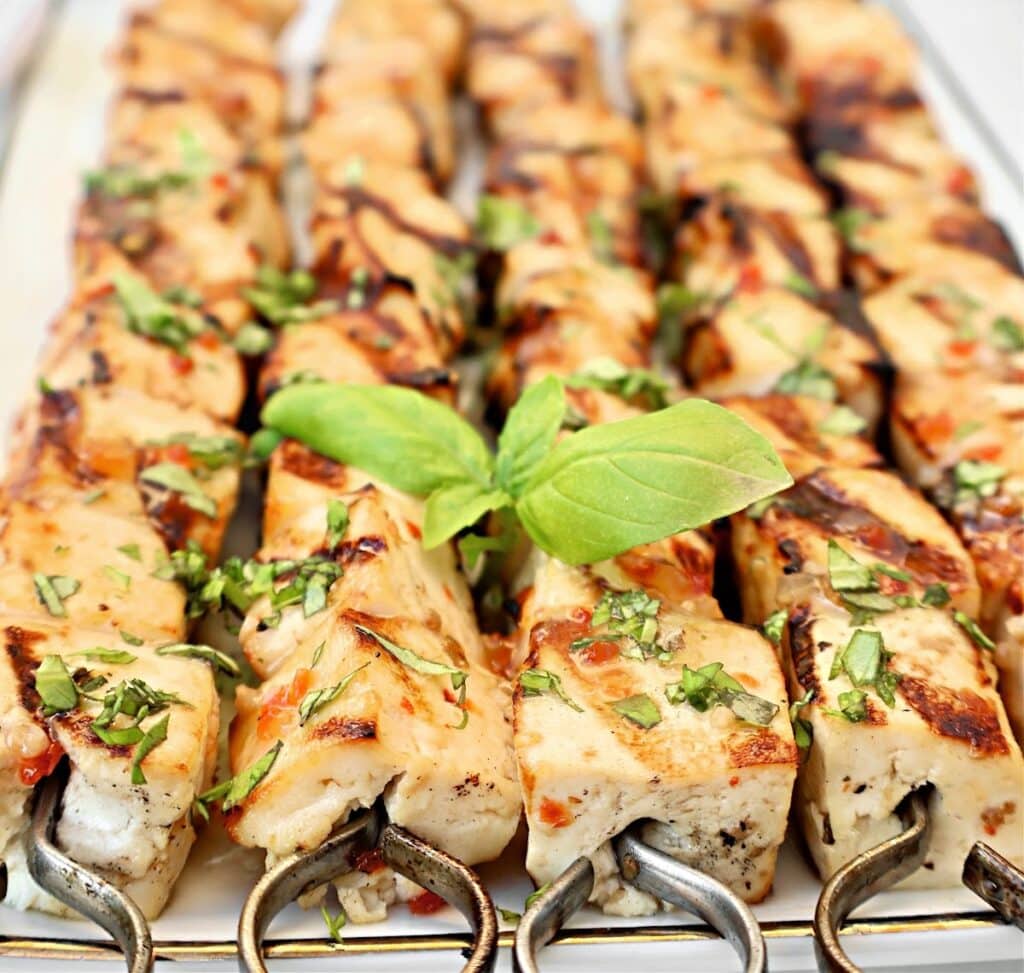 Grilled tofu skewers topped with chopped herbs are arranged on a platter, garnished with fresh basil leaves in the center. The meat is golden brown with visible grill marks.