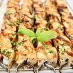 Grilled tofu skewers topped with chopped herbs are arranged on a platter, garnished with fresh basil leaves in the center. The meat is golden brown with visible grill marks.
