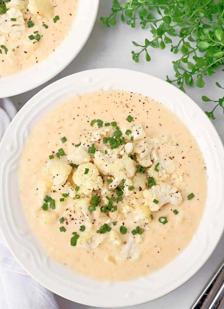 A bowl of creamy cauliflower soup topped with chopped chives and black pepper, served on a white plate with a sprig of fresh greens nearby.