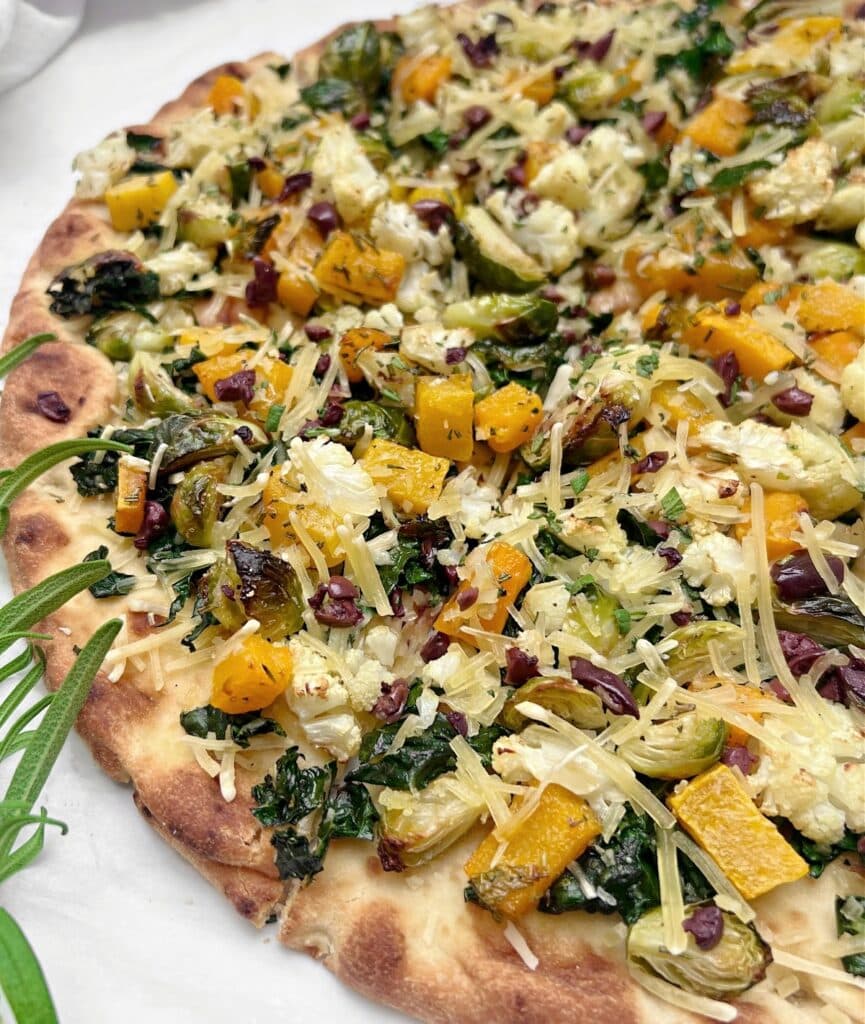 A close-up of a flatbread topped with roasted vegetables, including yellow squash, brussels sprouts, greens, grated cheese, and herbs on a crispy golden crust. A sprig of fresh rosemary is visible on the side.