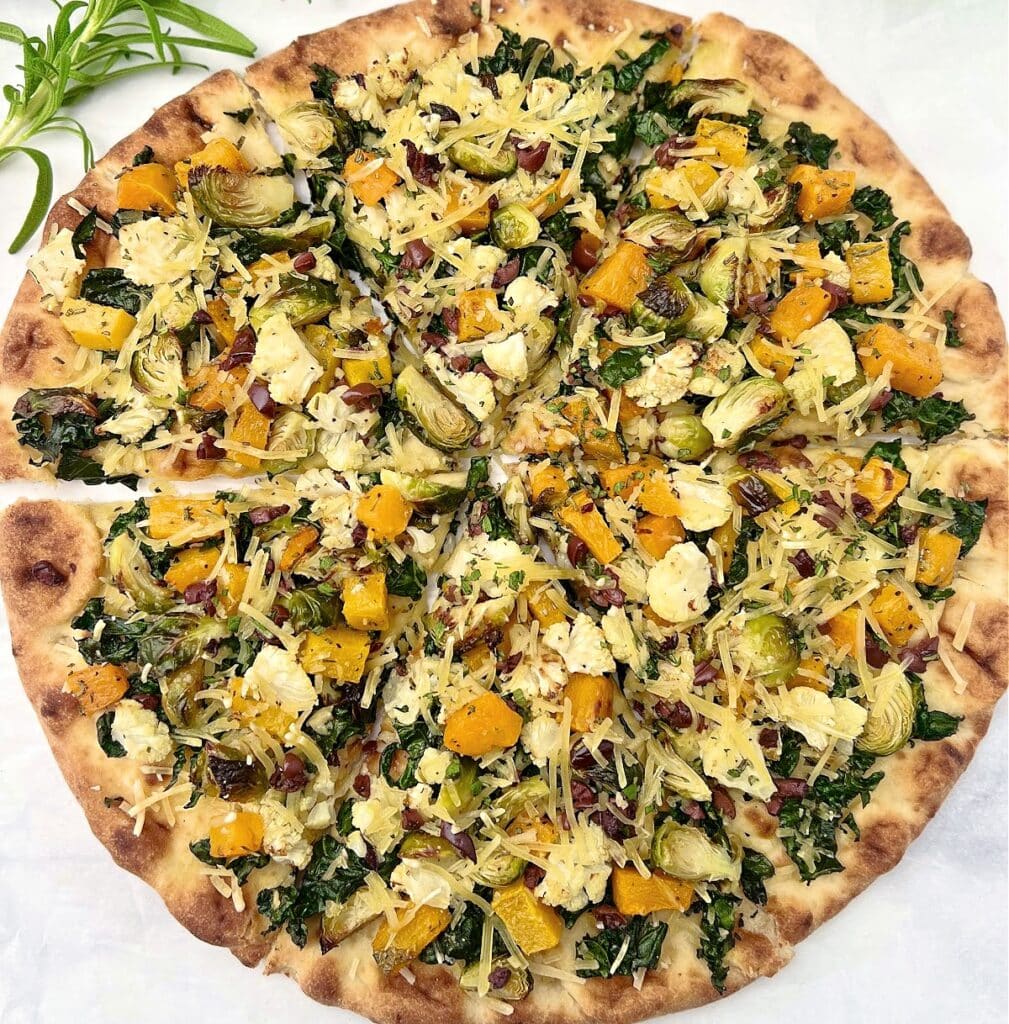 A thin-crust pizza topped with chopped kale, Brussels sprouts, roasted butternut squash, shredded cheese, and small bits of what appears to be bacon, sliced into eight pieces on parchment paper.
