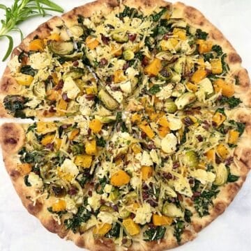 A thin-crust pizza topped with chopped kale, Brussels sprouts, roasted butternut squash, shredded cheese, and small bits of what appears to be bacon, sliced into eight pieces on parchment paper.
