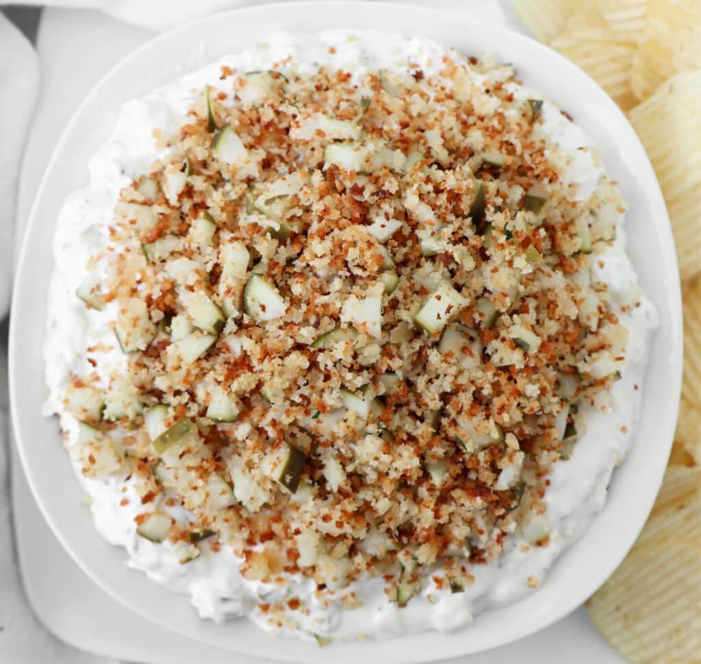 A white bowl filled with a creamy dip topped with chopped pickles and crispy browned breadcrumbs, surrounded by potato chips on the side.