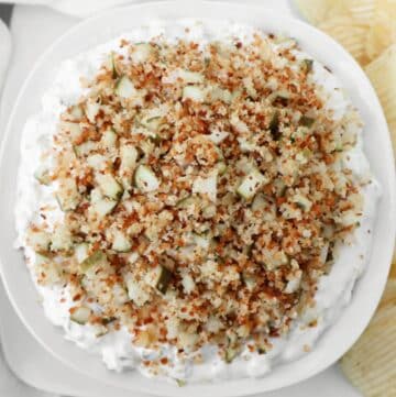 A white bowl filled with a creamy dip topped with chopped pickles and crispy browned breadcrumbs, surrounded by potato chips on the side.