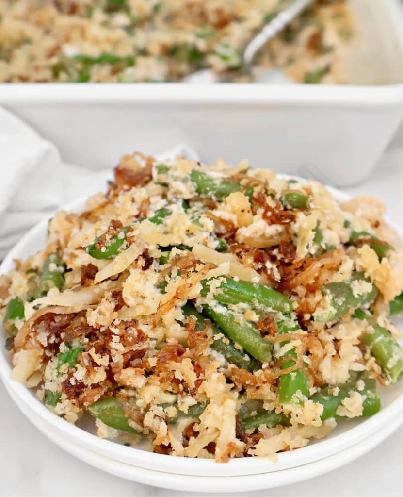A bowl of green bean casserole topped with crispy fried onions and breadcrumbs, with a baking dish of the same casserole in the background.