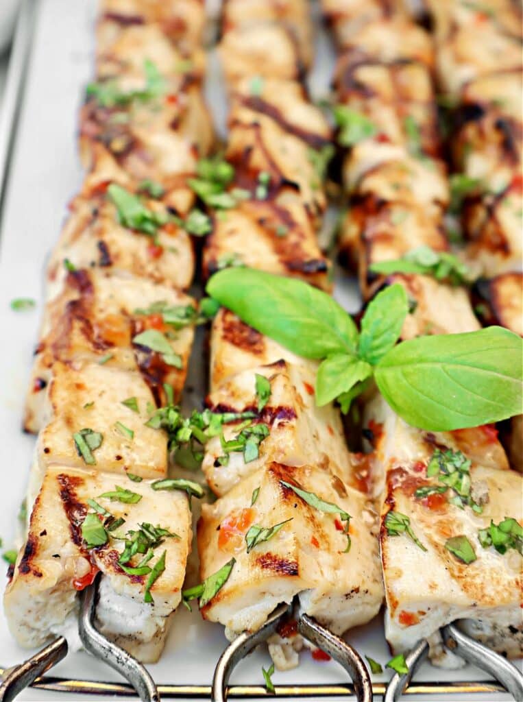 Grilled tofu skewers garnished with chopped herbs and a fresh basil sprig, arranged neatly on a white platter. The chicken has appetizing grill marks and a golden, slightly charred appearance.