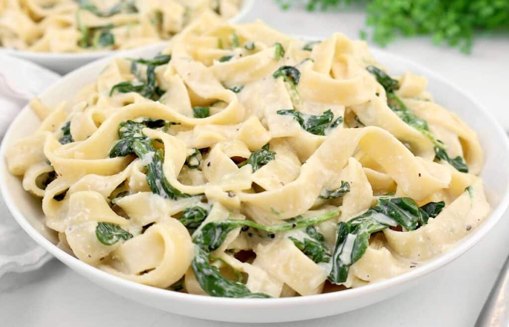 A white bowl filled with creamy fettuccine pasta mixed with wilted spinach, garnished with a light sauce. The background has a blurred hint of greenery.