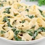 A white bowl filled with creamy fettuccine pasta mixed with wilted spinach, garnished with a light sauce. The background has a blurred hint of greenery.