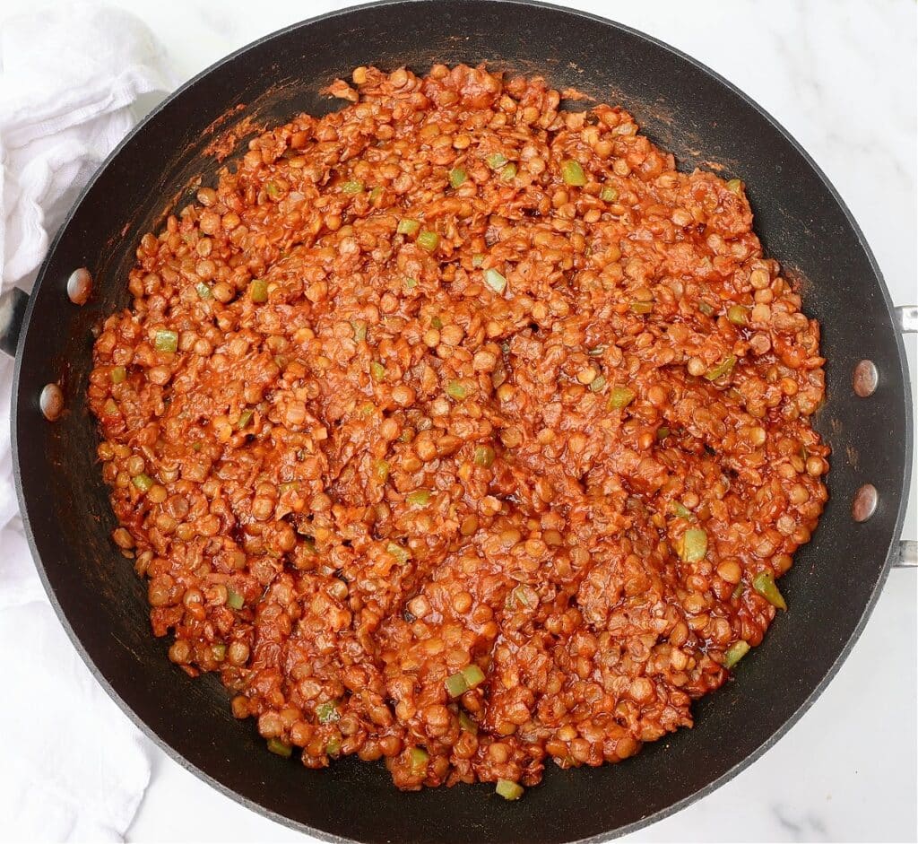 A large black skillet filled with cooked lentils in a thick, red, tomato-based sauce with visible diced green vegetables, sitting on a white marble surface.