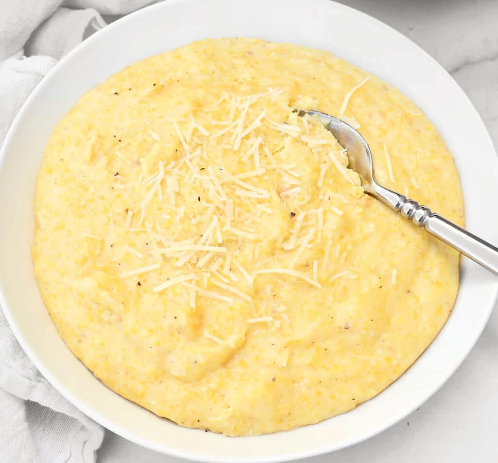 A white bowl filled with creamy yellow polenta, topped with shredded cheese, with a silver spoon resting in the dish.