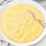 A white bowl filled with creamy yellow polenta, topped with shredded cheese, with a silver spoon resting in the dish.