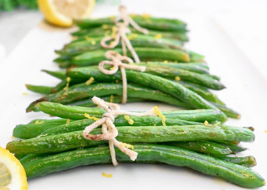 Neatly tied green bean bundles with twine, garnished with lemon zest, are arranged on a white plate. A slice of lemon is visible in the background.