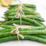 Neatly tied green bean bundles with twine, garnished with lemon zest, are arranged on a white plate. A slice of lemon is visible in the background.