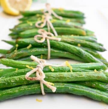 Neatly tied green bean bundles with twine, garnished with lemon zest, are arranged on a white plate. A slice of lemon is visible in the background.