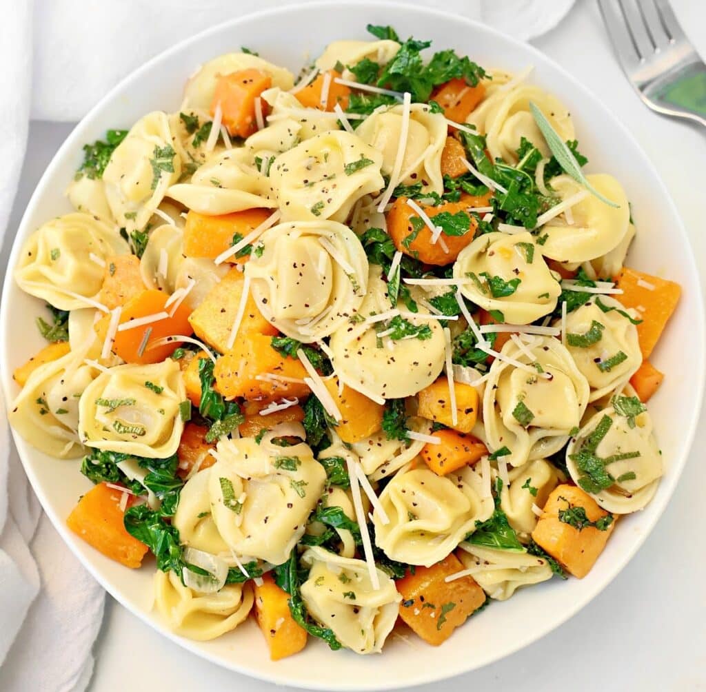 A bowl of tortellini with butternut squash, chopped kale, fresh herbs, and grated parmesan cheese. A fork and white napkin sit beside this inviting pasta dish.