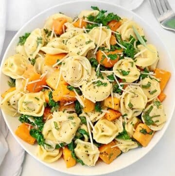 A bowl of tortellini with butternut squash, chopped kale, fresh herbs, and grated parmesan cheese. A fork and white napkin sit beside this inviting pasta dish.