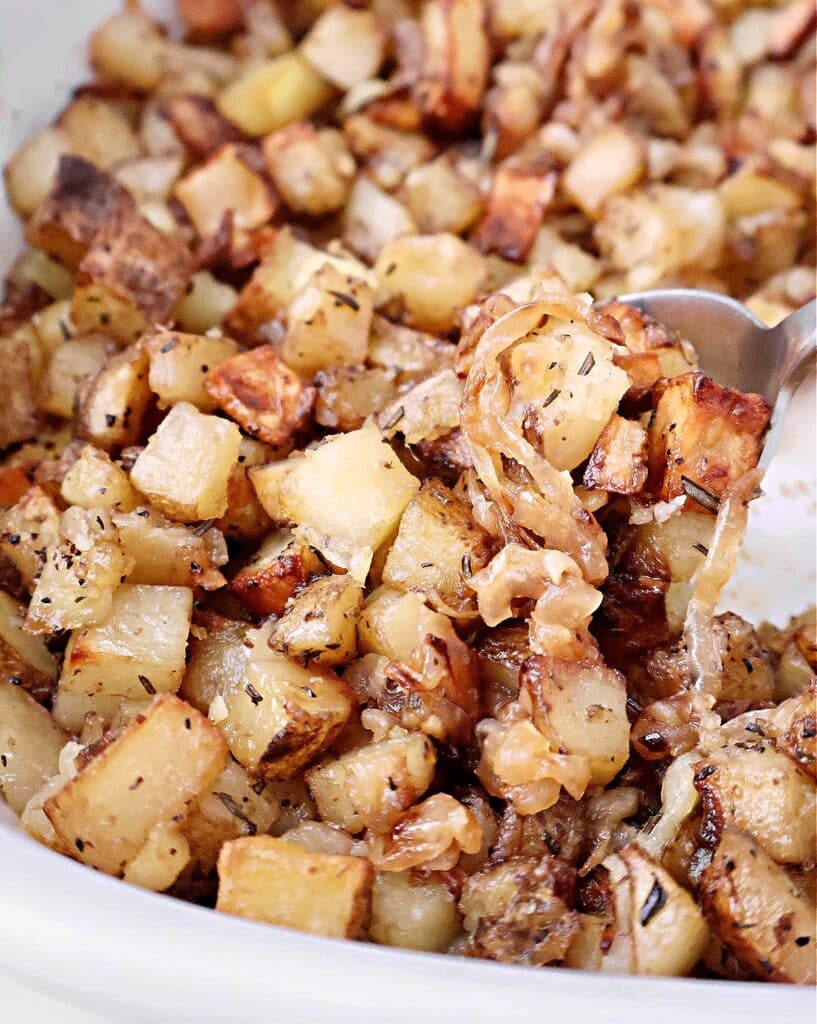 Close-up of roasted breakfast potatoes mixed with caramelized onions and herbs, served in a dish with a metal spoon. The crispy, golden brown potatoes look perfect for a hearty morning meal.