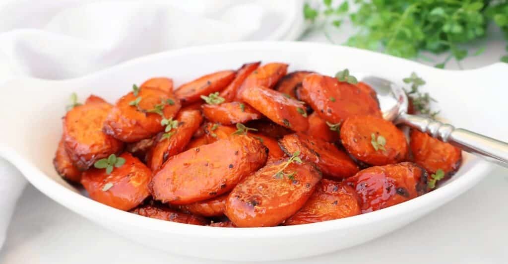 A white oval dish filled with balsamic roasted carrots, their glazed slices garnished with fresh green herbs; a silver spoon rests on the right side of the dish.