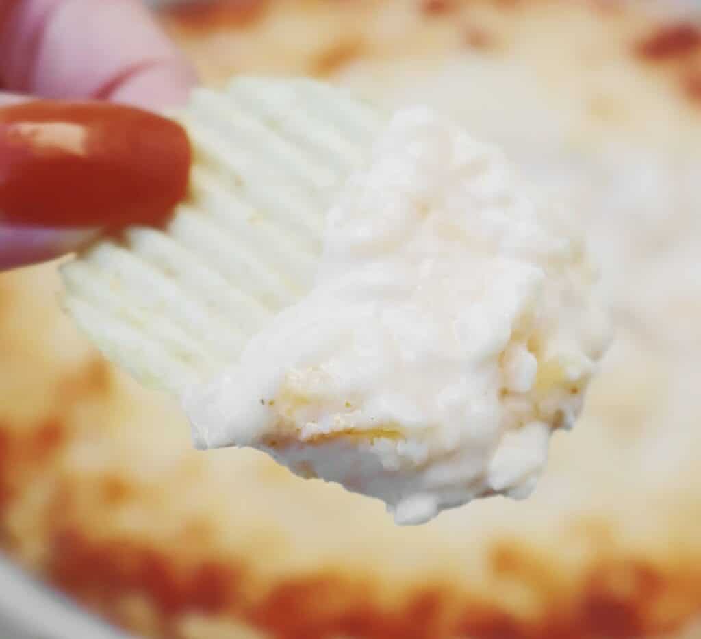 A hand with painted nails holds a ridged potato chip dipped in a creamy, white cheese dip, with more dip visible blurred in the background.
