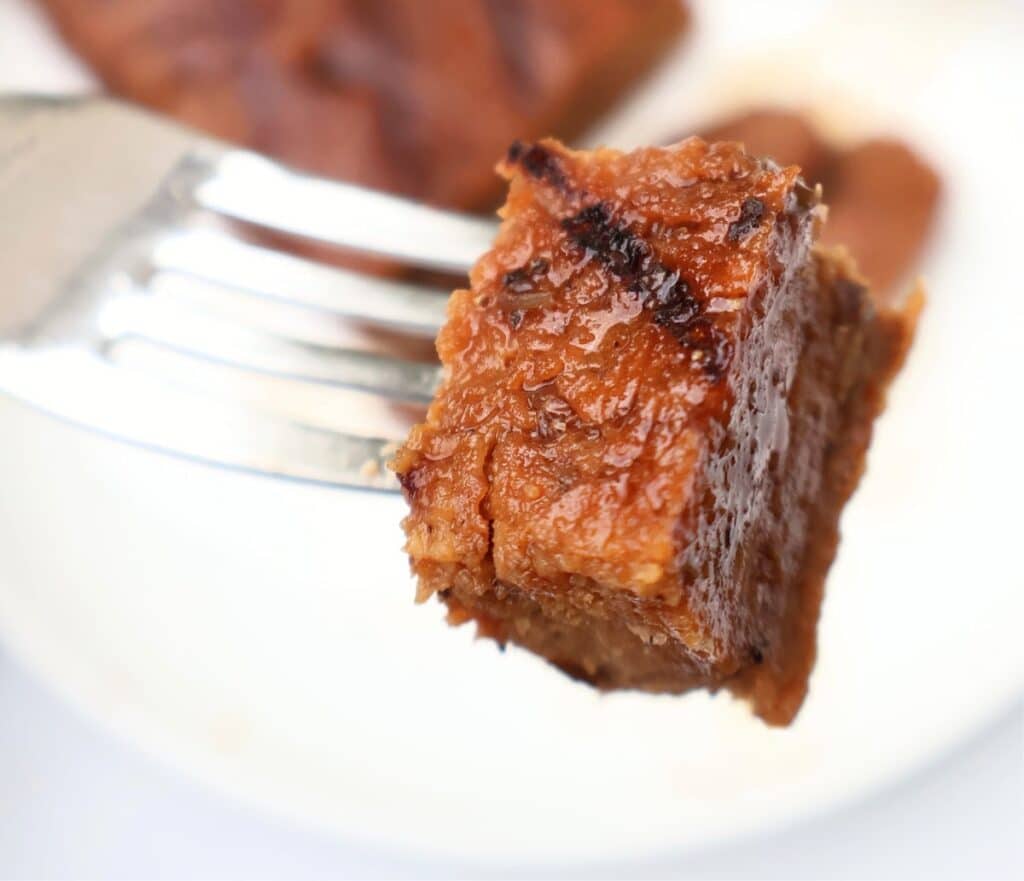 A close-up of a fork holding a juicy, browned piece of cooked vegan meat with a slightly crispy exterior, against a blurred white plate in the background.