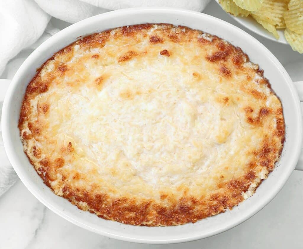A white baking dish filled with golden-brown, bubbly, baked dip, surrounded by ridged potato chips and white napkins on a marble surface.
