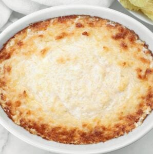 A baked cheesy dip in a round white dish, golden and slightly browned on top, with a creamy texture and crispy edges, placed on a white surface.