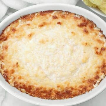 A baked cheesy dip in a round white dish, golden and slightly browned on top, with a creamy texture and crispy edges, placed on a white surface.