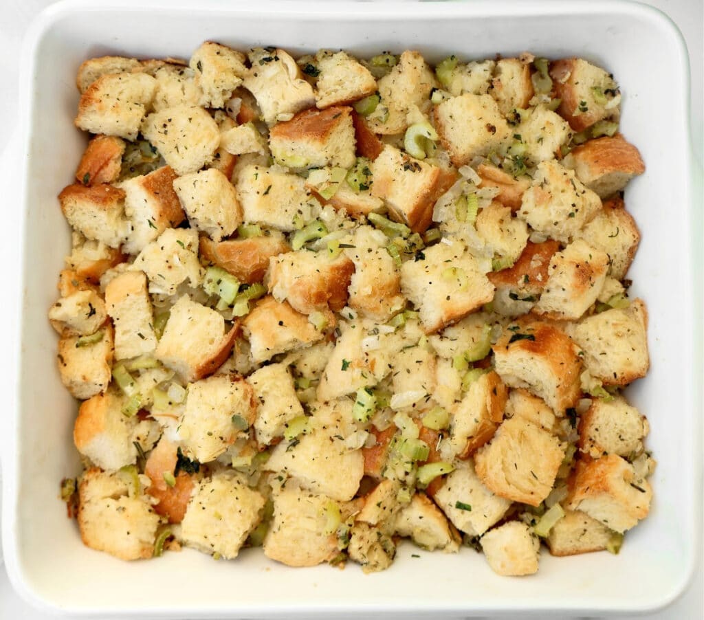 A white baking dish filled with cubed bread pieces mixed with chopped celery, herbs, and seasonings, resembling a classic stuffing or dressing before baking.