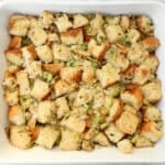 A white baking dish filled with cubed bread pieces mixed with chopped celery, herbs, and seasonings, resembling a classic stuffing or dressing before baking.