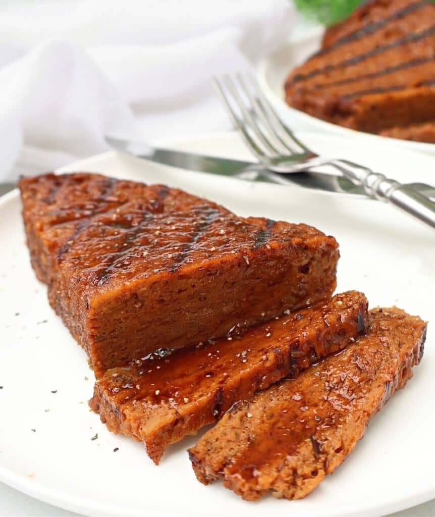 Sliced, grilled seitan steak on a white plate, garnished with herbs and drizzled with sauce. A fork and knife rest behind the steak, with another steak in the background.