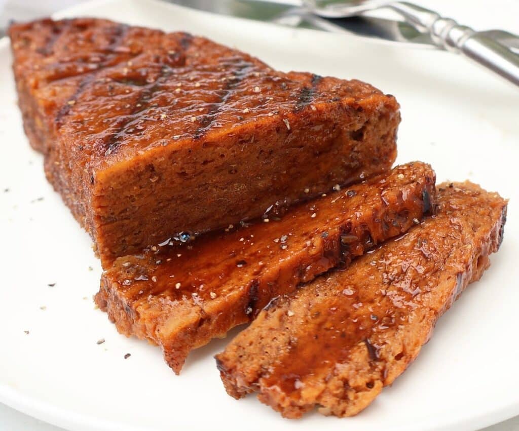 Sliced vegan steak with a shiny glaze sits on a white plate, showing a moist, tender texture. A fork and knife are visible in the background.
