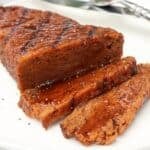Sliced vegan steak with a shiny glaze sits on a white plate, showing a moist, tender texture. A fork and knife are visible in the background.