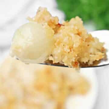 A close-up of a fork holding a bite of cheesy creamed onions topped with golden, crispy breadcrumbs. The background is blurred, showcasing a plate of creamy pearl onions and some green garnish.
